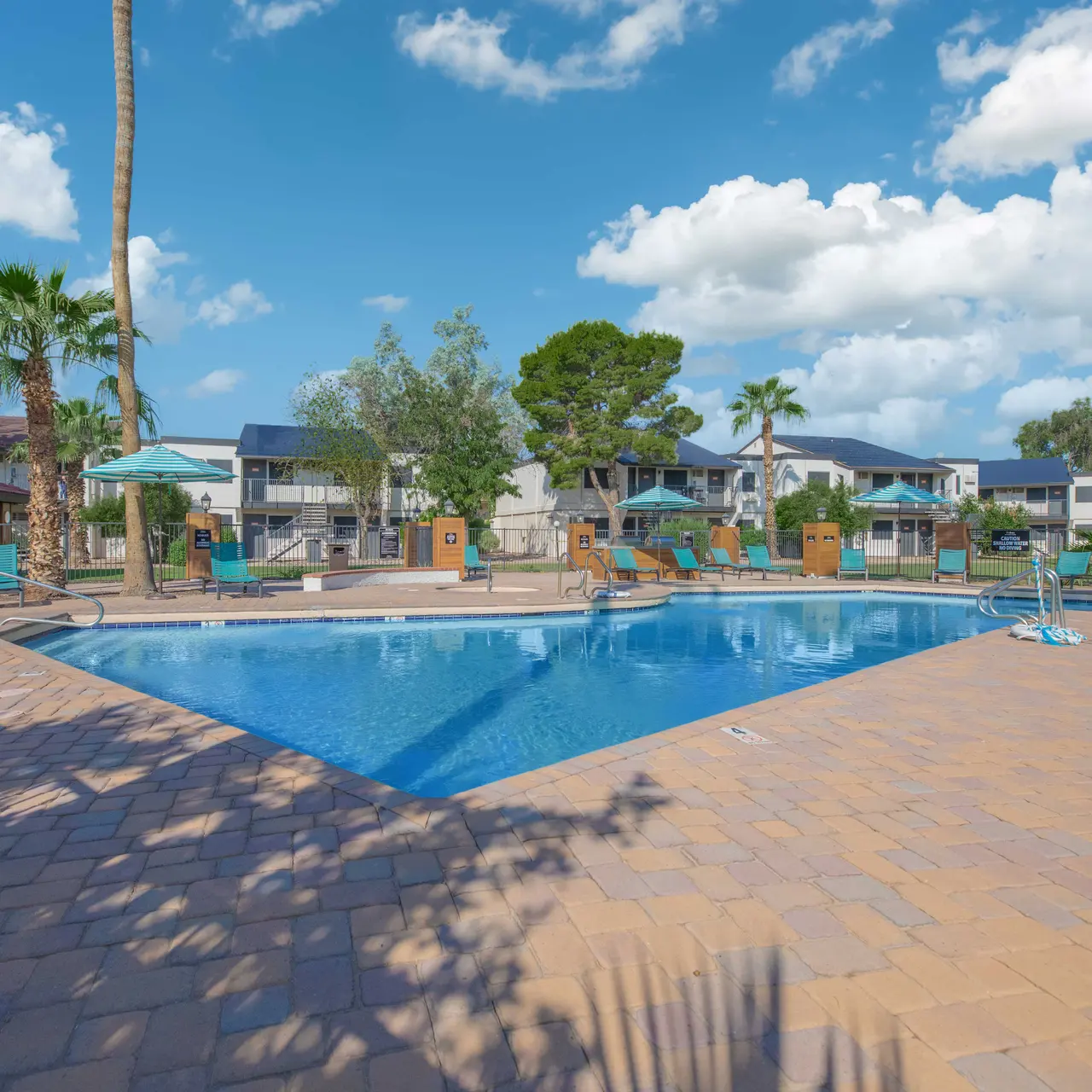 A spacious swimming pool area with lounge chairs, umbrellas, and surrounding palm trees under a bright blue sky with clouds.