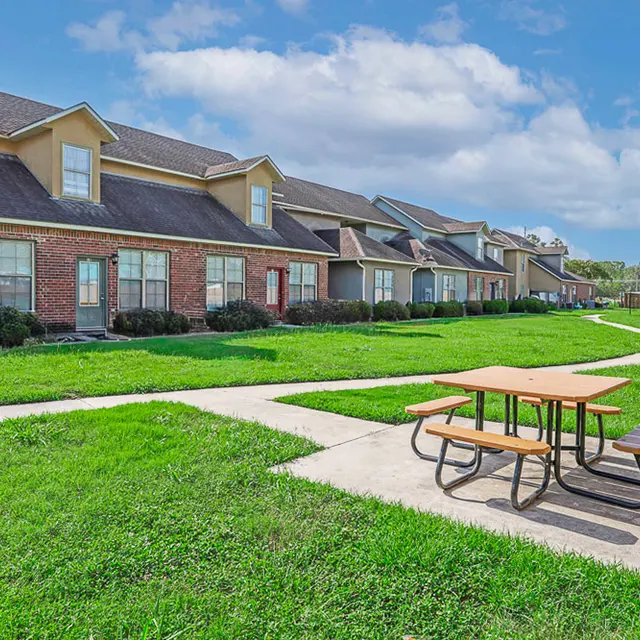 A well-maintained apartment complex featuring a grassy area with a picnic table, surrounded by several residential buildings under a blue sky.