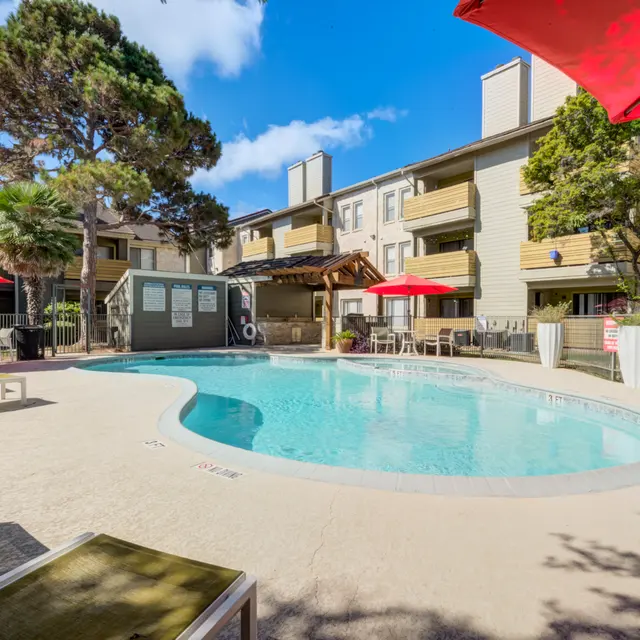A clear blue swimming pool surrounded by lounge chairs and red umbrellas in an apartment complex.