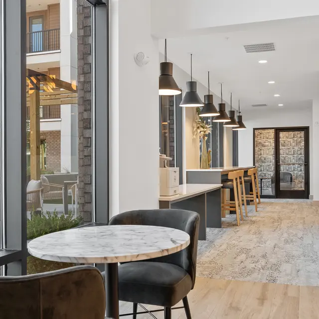 A modern café interior featuring a round marble table and black chairs. The space is filled with natural light from large windows that overlook an outdoor seating area. White walls and wooden accents create a bright and inviting ambiance.
