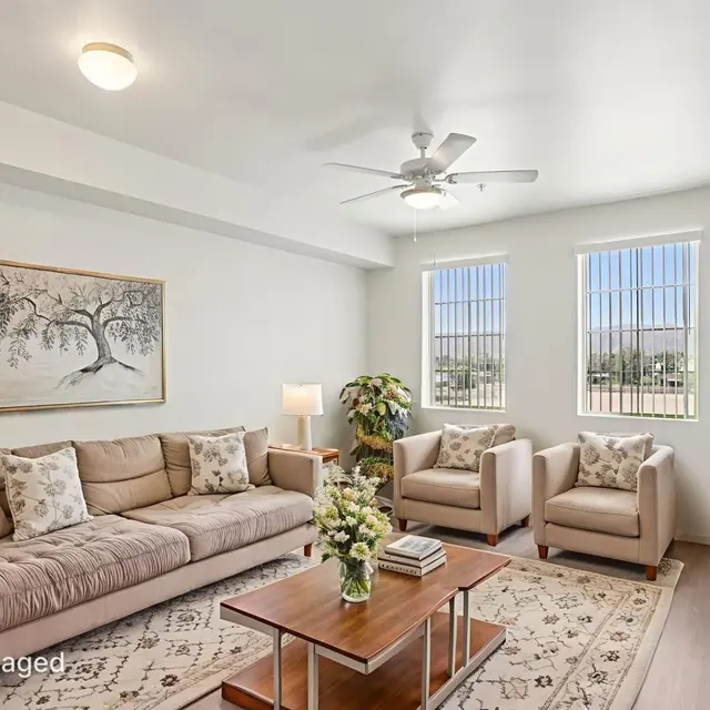 Modern Living Room Design A cozy living room featuring a beige sofa, two armchairs, a wooden coffee table, and a decorative rug. The room has large windows letting in natural light, with green plants and wall art visible.