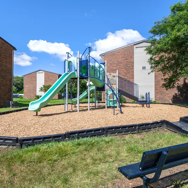 A playground featuring a slide and climbing structure, surrounded by grass and a bench, with apartment buildings in the background under a clear blue sky.