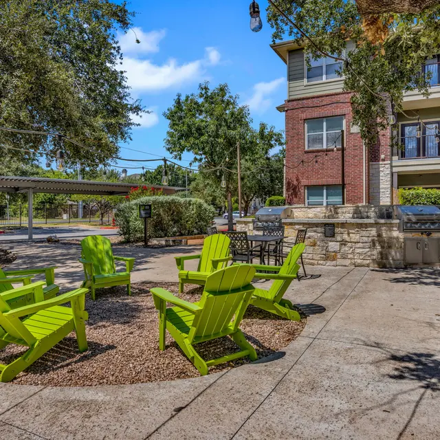 An outdoor seating area featuring several bright green Adirondack chairs arranged in a circular layout on a stone surface, with a barbeque grill nearby and a multi-story brick building in the background.