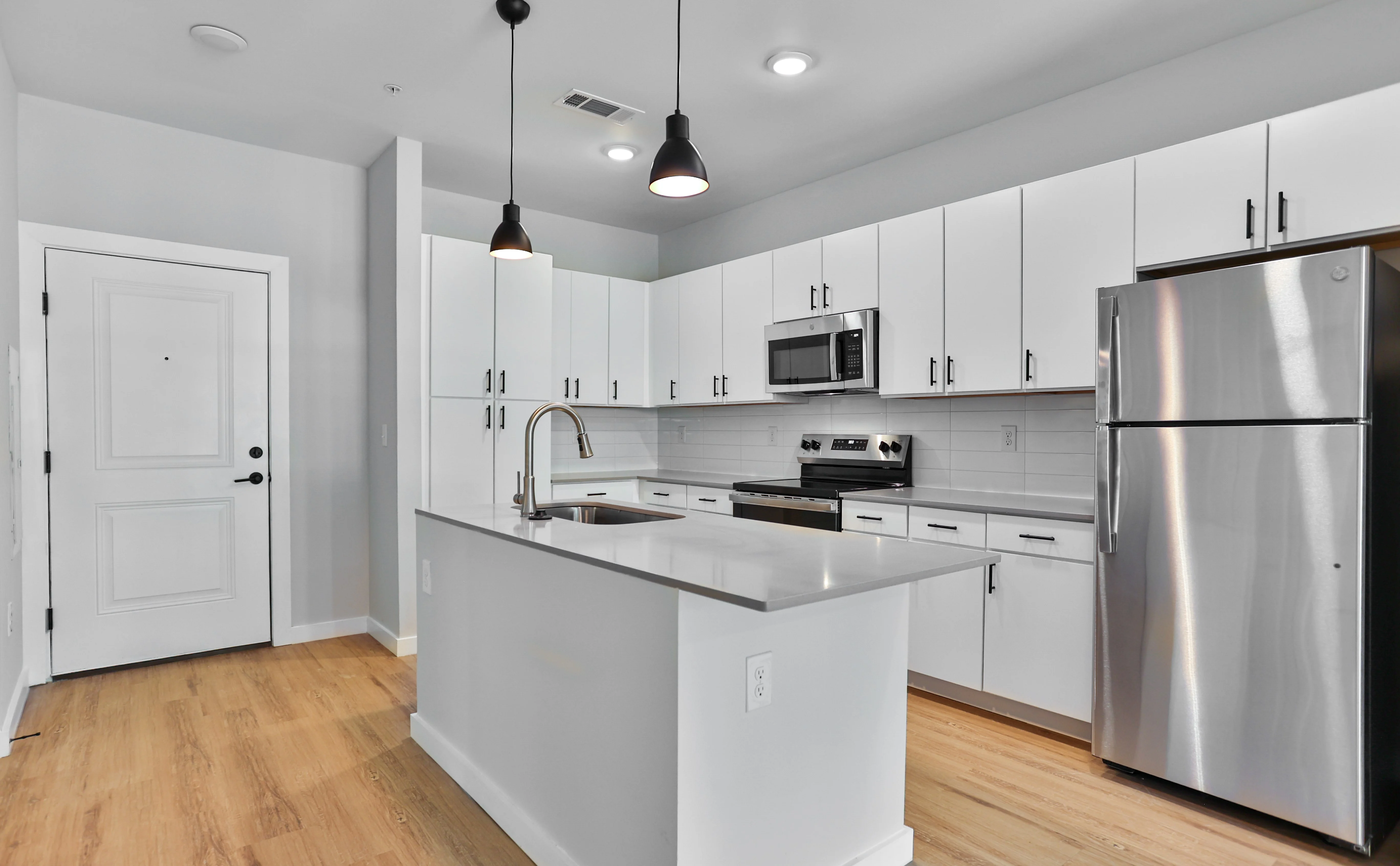 A modern kitchen featuring white cabinets, stainless steel appliances, and a large island with a sink. The room has wood flooring and bright lighting from pendant lights.