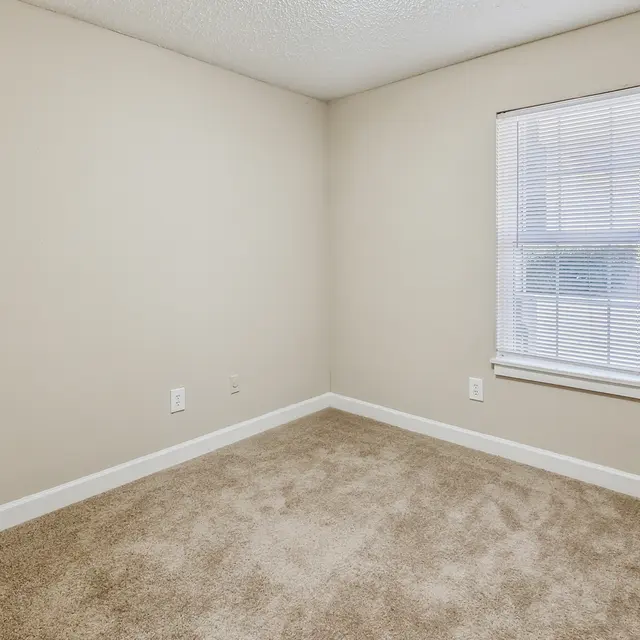 A vacant room with light beige walls and carpet, featuring a single window with blinds, illuminating the space.