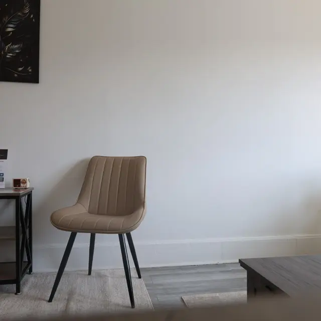 A minimalist living room featuring a light-colored wall, a beige chair, a small black side table with magazines, and a green plant in the corner.