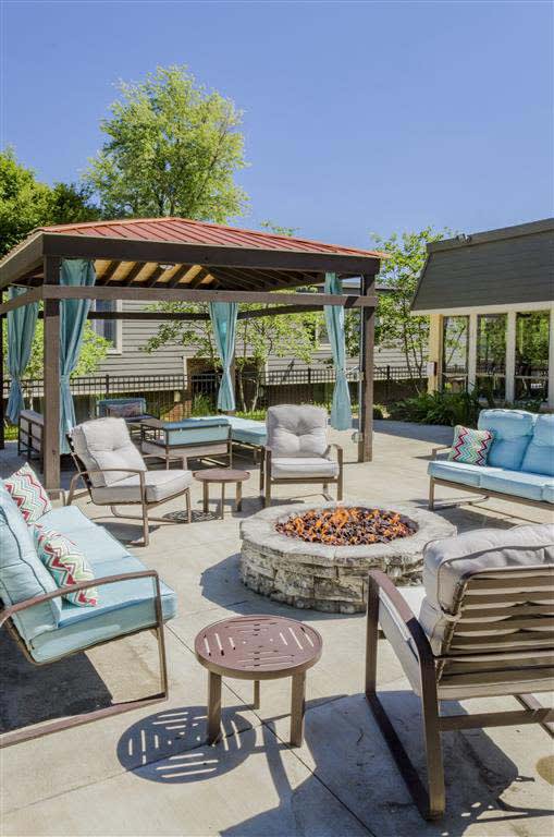 A cozy outdoor seating area featuring a circular stone fire pit surrounded by cushioned chairs and a gazebo with draped curtains.