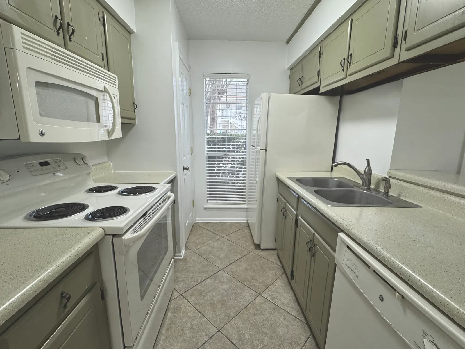 A compact kitchen featuring green cabinetry, white appliances including a stove, microwave, refrigerator, and dishwasher. The counter is light-colored with a sink visible. A window with blinds provides natural light.