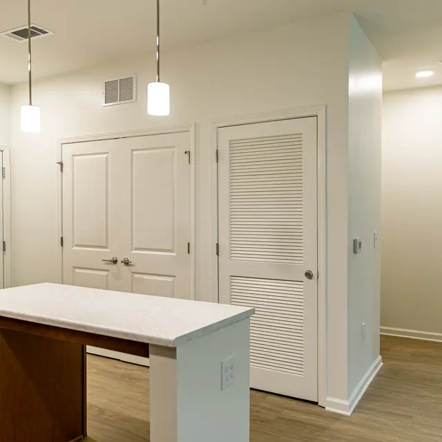 A modern entryway featuring two doors, one with a peephole and the other with horizontal slats. A small kitchen island with a white countertop and dark wood base is in the foreground, illuminated by pendant lights. The walls are painted white and the flooring is a light wood finish.
