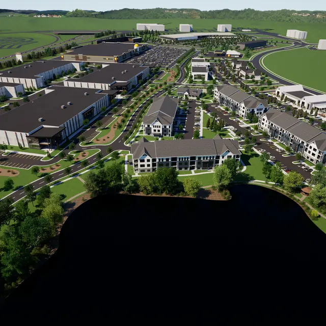 Aerial view of a large modern development with multiple buildings, roads, and green spaces, adjacent to a body of water.
