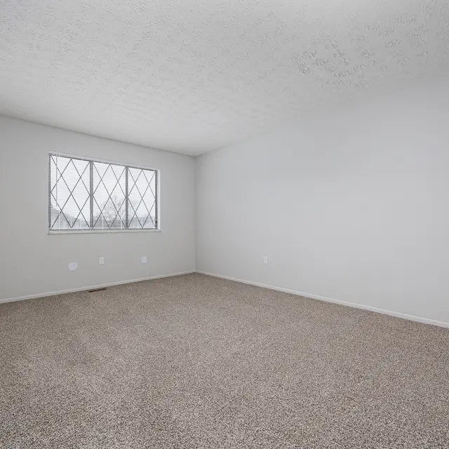 An empty room with light gray walls and a textured ceiling, featuring a large window with a diamond-patterned grille. The floor is covered in beige carpet, and there are no furnishings present.