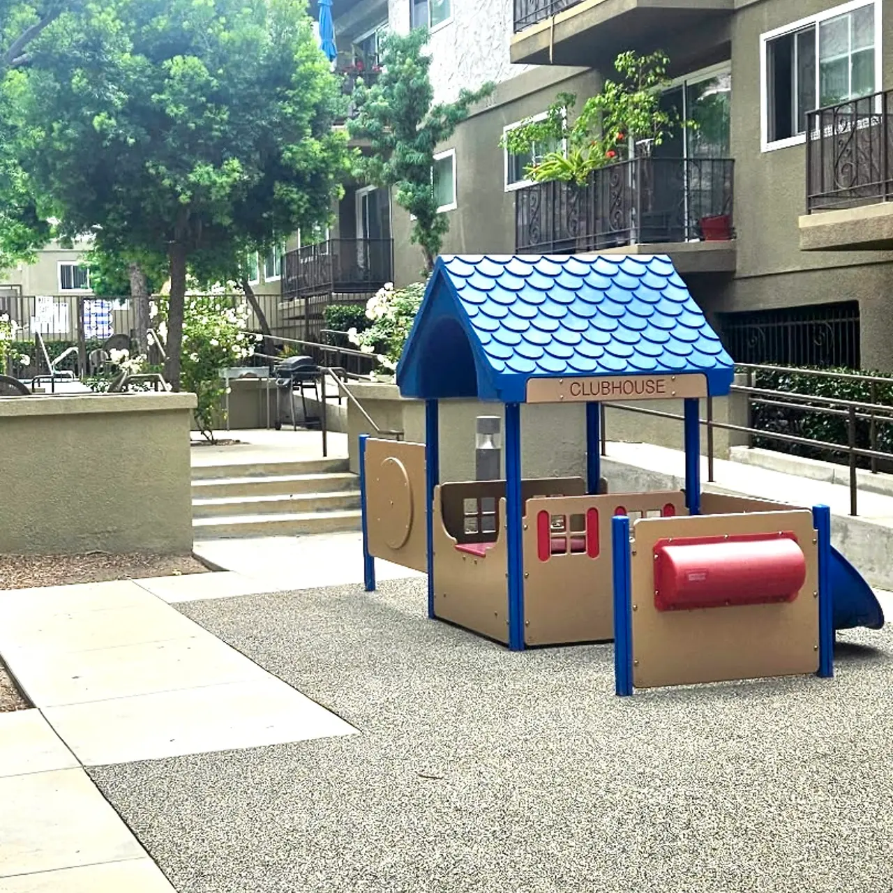 A colorful children's playhouse in a playground surrounded by concrete walkways and plants with apartment buildings in the background.