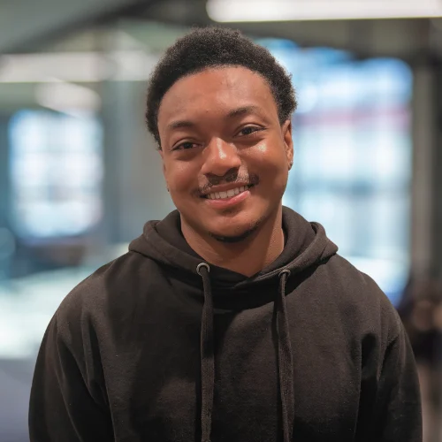 A young man smiling, wearing a black hoodie, with a soft-focus background of blurred lights.