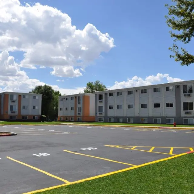 A view of a multi-unit apartment complex with three buildings, surrounded by a parking lot and green grass under a blue sky with fluffy clouds.
