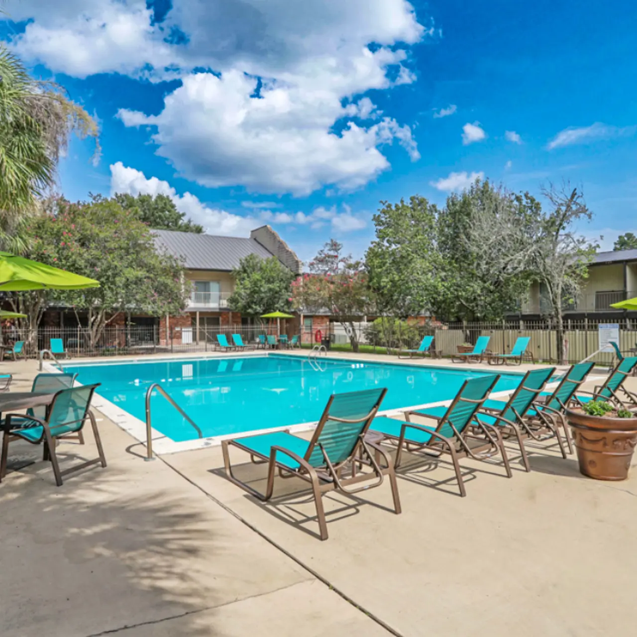 A sunny pool area surrounded by lounge chairs and green umbrellas, with a clear blue sky and nearby trees.