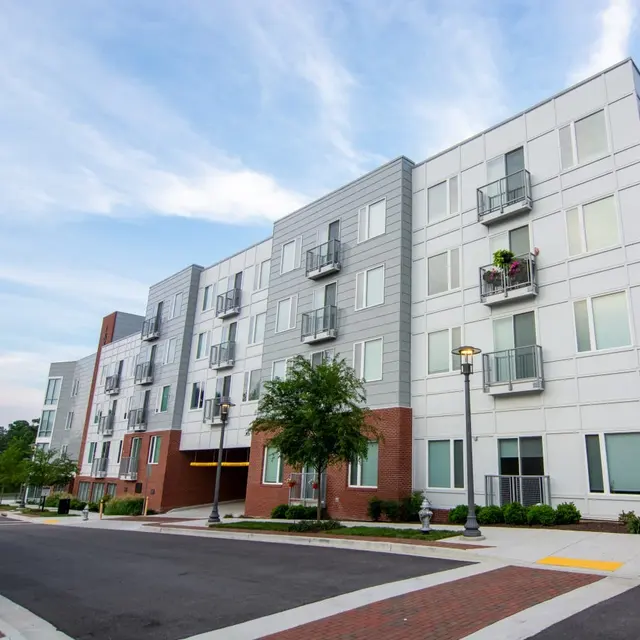 A modern apartment building with multiple floors, featuring balconies and large windows, surrounded by green landscaping and paved walkways.