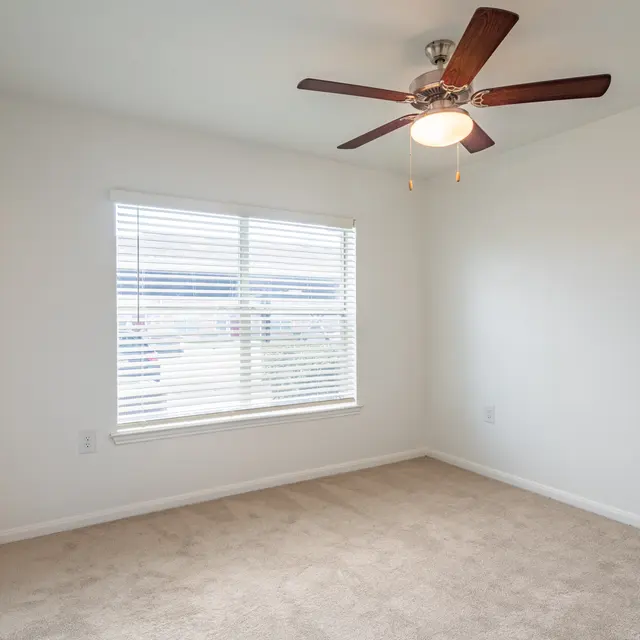A bright and empty bedroom with a light carpet, a ceiling fan, and a window with blinds.