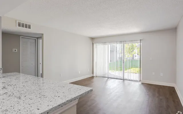 Modern Apartment Living Room An empty modern apartment interior featuring a kitchen area with a granite countertop, a spacious living area, and a wall of sliding glass doors leading to a green outdoor space. The walls are painted in neutral colors with a textured ceiling and wood-like flooring.
