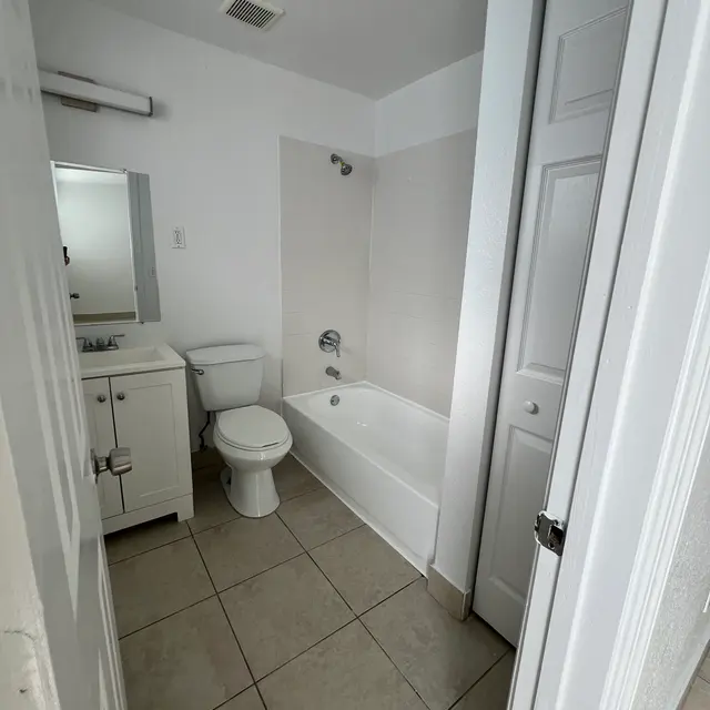 A clean, modern bathroom featuring a bathtub, toilet, sink, and mirror, with light-colored tiles and a door to the hallway.