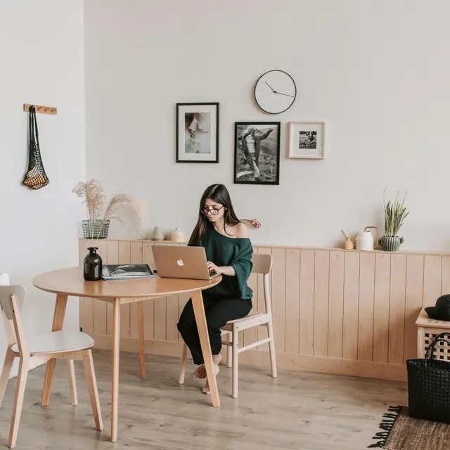 A woman sitting at a round wooden table, working on a laptop in a modern home office setting decorated with minimalist artwork and plants.
