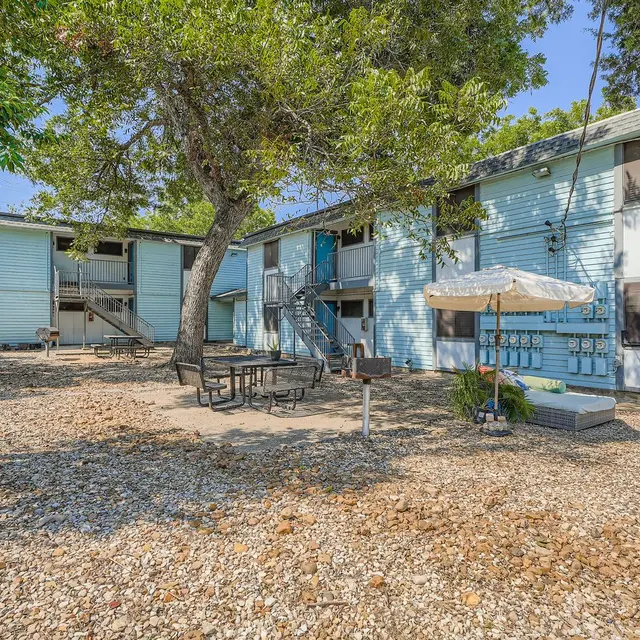 Outdoor area of a blue apartment complex featuring a picnic table, shaded seating, and a tree.