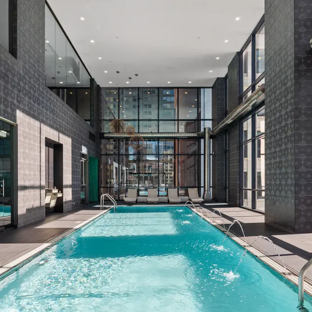 A modern indoor swimming pool surrounded by large glass windows, with sleek dark walls and a clean design. Sunlight brightens the space.