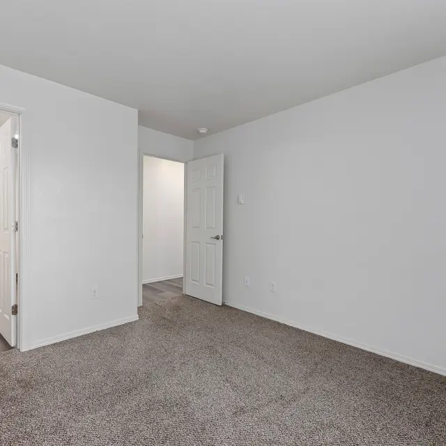 Empty Room with Neutral Decor An empty room with light gray walls and carpeted flooring, featuring a doorway leading to another room and a door closed on the right.
