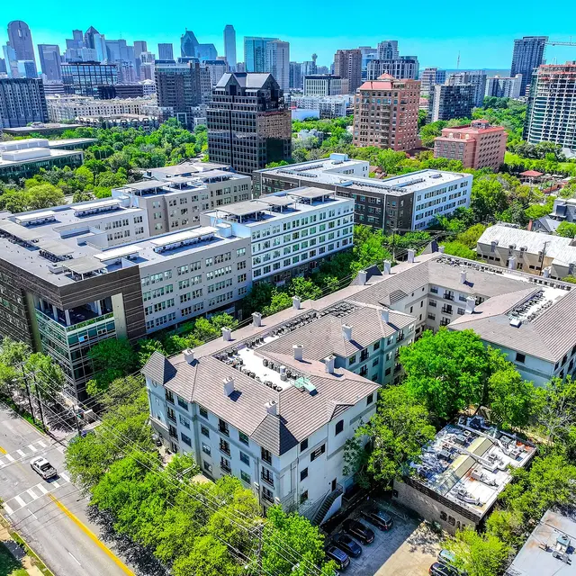 Aerial view of a vibrant urban landscape featuring multiple modern buildings, lush green trees, and a clear blue sky. The skyline of downtown Dallas is visible in the background.
