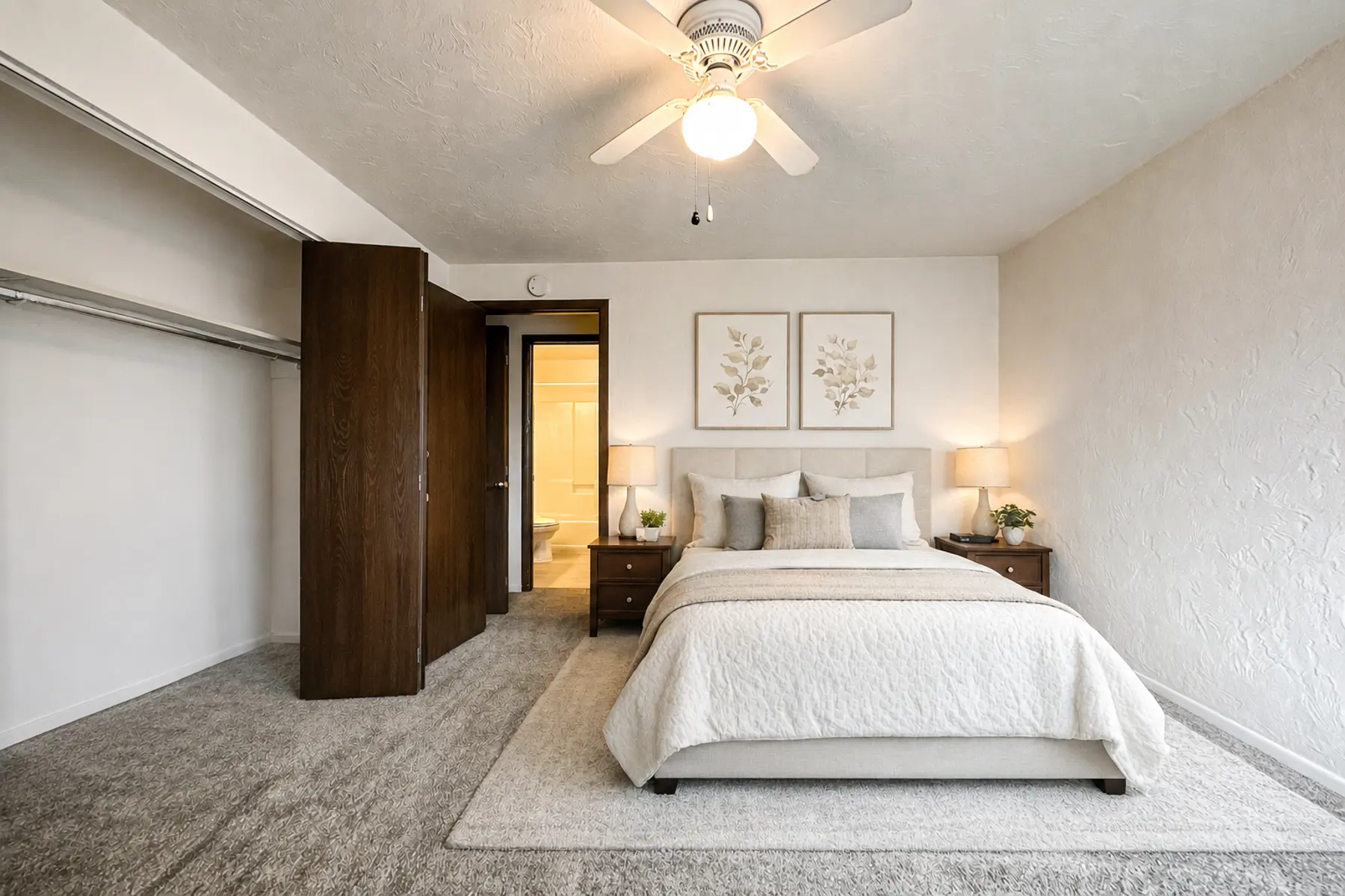 A bright and modern bedroom featuring a large bed with light bedding, nightstands on either side, and simple wall art. There's a closet with open doors and warm lighting in the room.