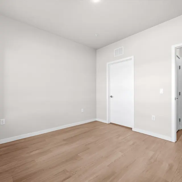 A spacious, empty room with light-colored walls and wooden flooring. A closed door leads to a bathroom area that is partially visible, with beige panels in the background. Natural light is coming from a window.