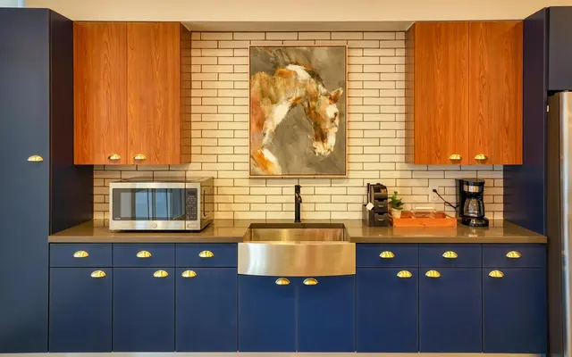 A modern kitchen featuring blue cabinets with gold handles, a stainless steel sink, a microwave, and a coffee maker. The decor includes a horse painting above a tiled backsplash.