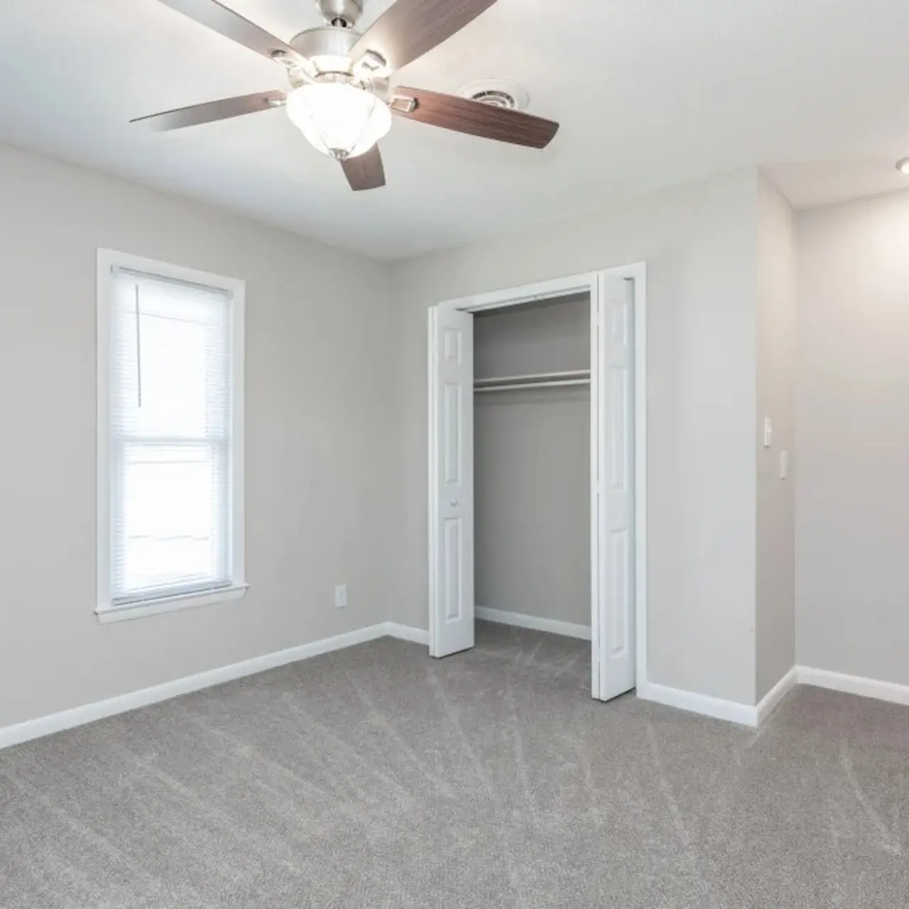 An empty room featuring light gray walls and carpet, a ceiling fan, and a closet with doors open. Two windows allow natural light.