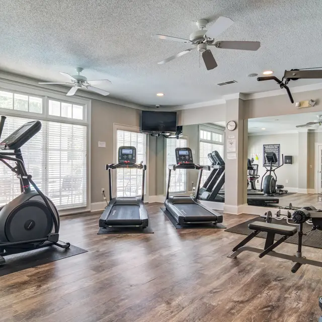 A well-lit modern gym interior with various fitness equipment including elliptical machines, treadmills, and a weightlifting station. Large windows allow natural light to fill the space.