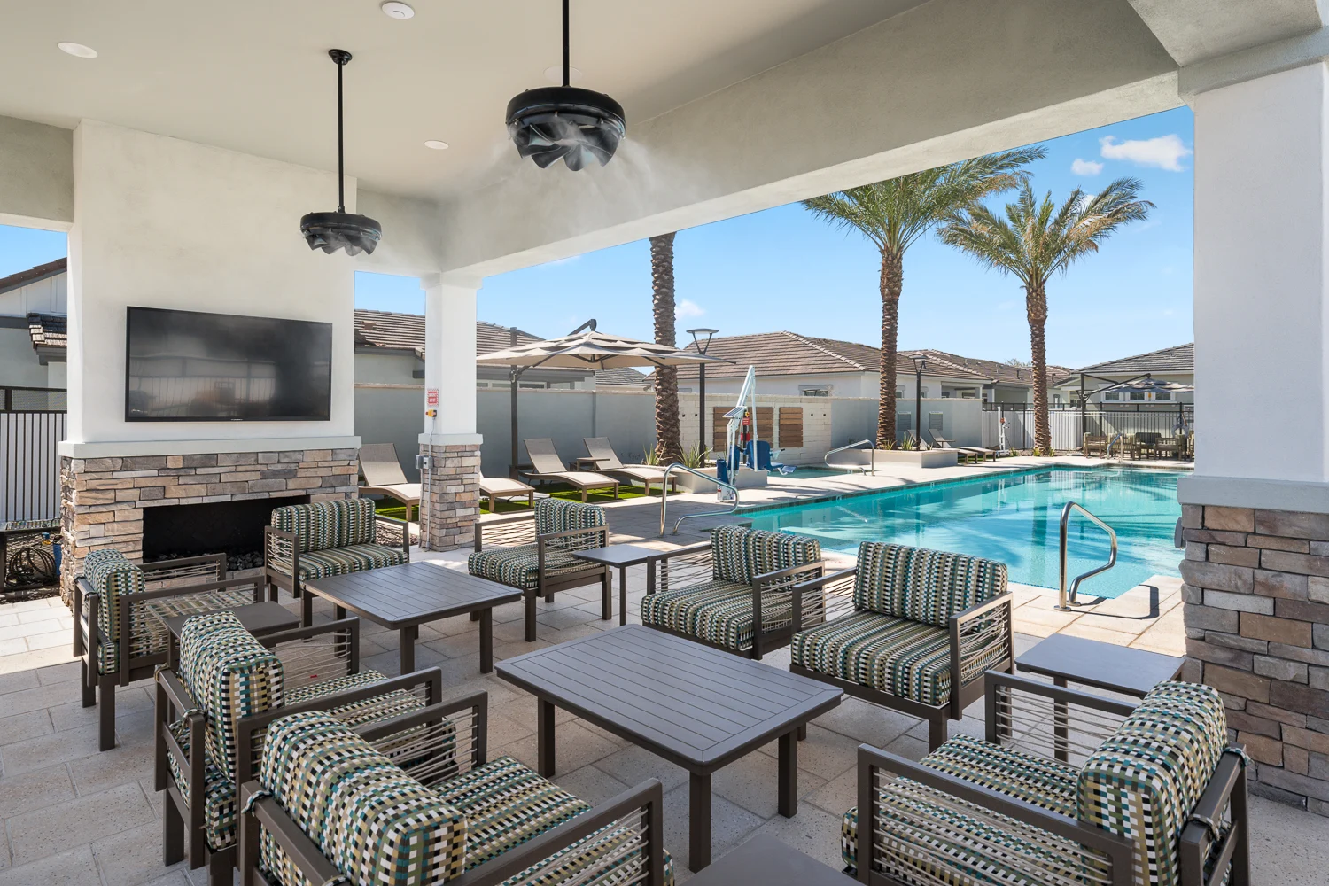 A modern outdoor lounge area next to a swimming pool, featuring several striped chairs and tables, a flat-screen TV mounted on the wall, and palm trees in the background under a clear blue sky.