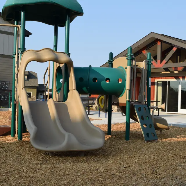 Playground Equipment in a Recreational Area A playground featuring a slide, climbing structure, and a play tunnel, set on a bed of wood chips with a building in the background.