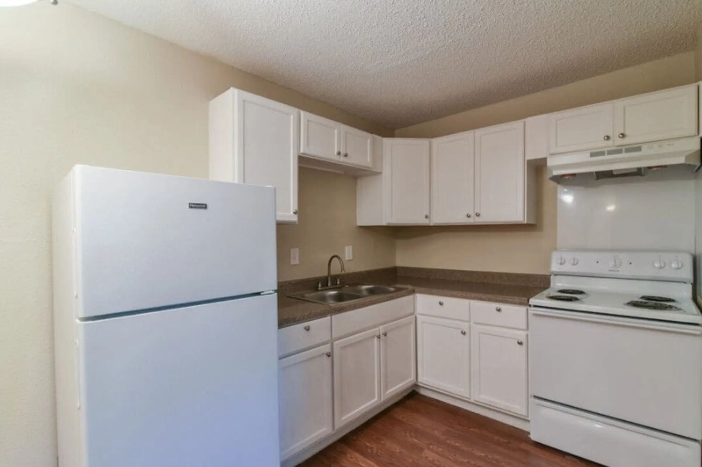 A simple kitchen with white cabinets, a refrigerator, and a stove, featuring a sink and countertop, with wooden flooring.