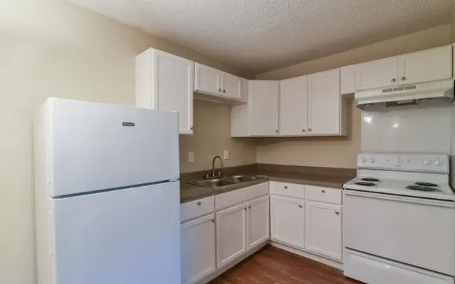 A simple kitchen with white cabinets, a refrigerator, and a stove, featuring a sink and countertop, with wooden flooring.