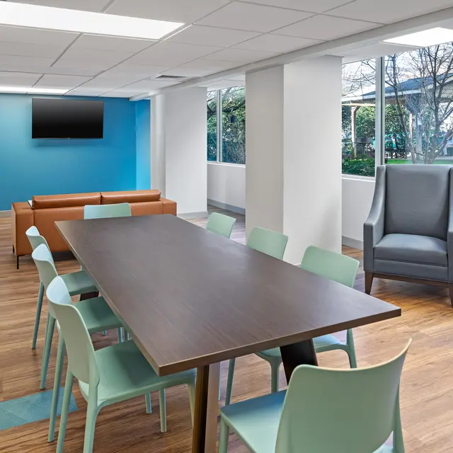 A spacious, modern meeting room with a long wooden table surrounded by colorful chairs, a brown leather couch, and large windows letting in natural light.