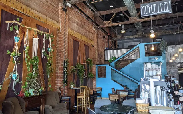 Interior of a coffee shop featuring exposed brick walls, plants, comfortable seating, and a staircase. A sign reads 'PICK UP HERE'.