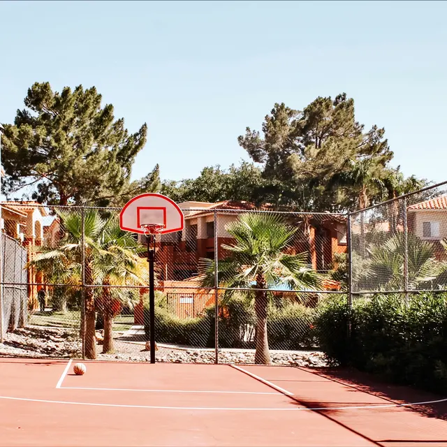 A basketball court surrounded by trees and residential buildings, featuring a hoop and a red-painted surface.