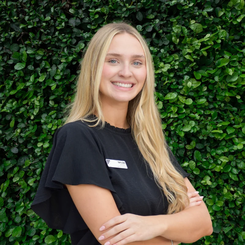 A smiling young woman with long blonde hair wearing a black top, standing outdoors in front of a lush green wall.