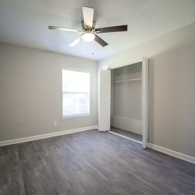 An empty bedroom with light gray walls and wooden flooring, featuring a ceiling fan and a window with blinds.
