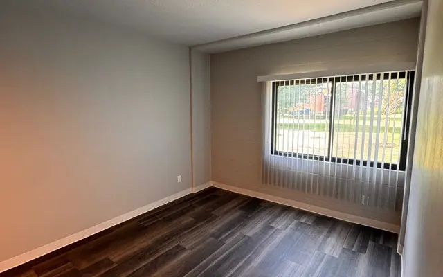 Empty Room Interior A spacious empty room with grey walls and dark wooden flooring. A large window with vertical blinds allows natural light to enter the room, and a view of greenery is visible outside.