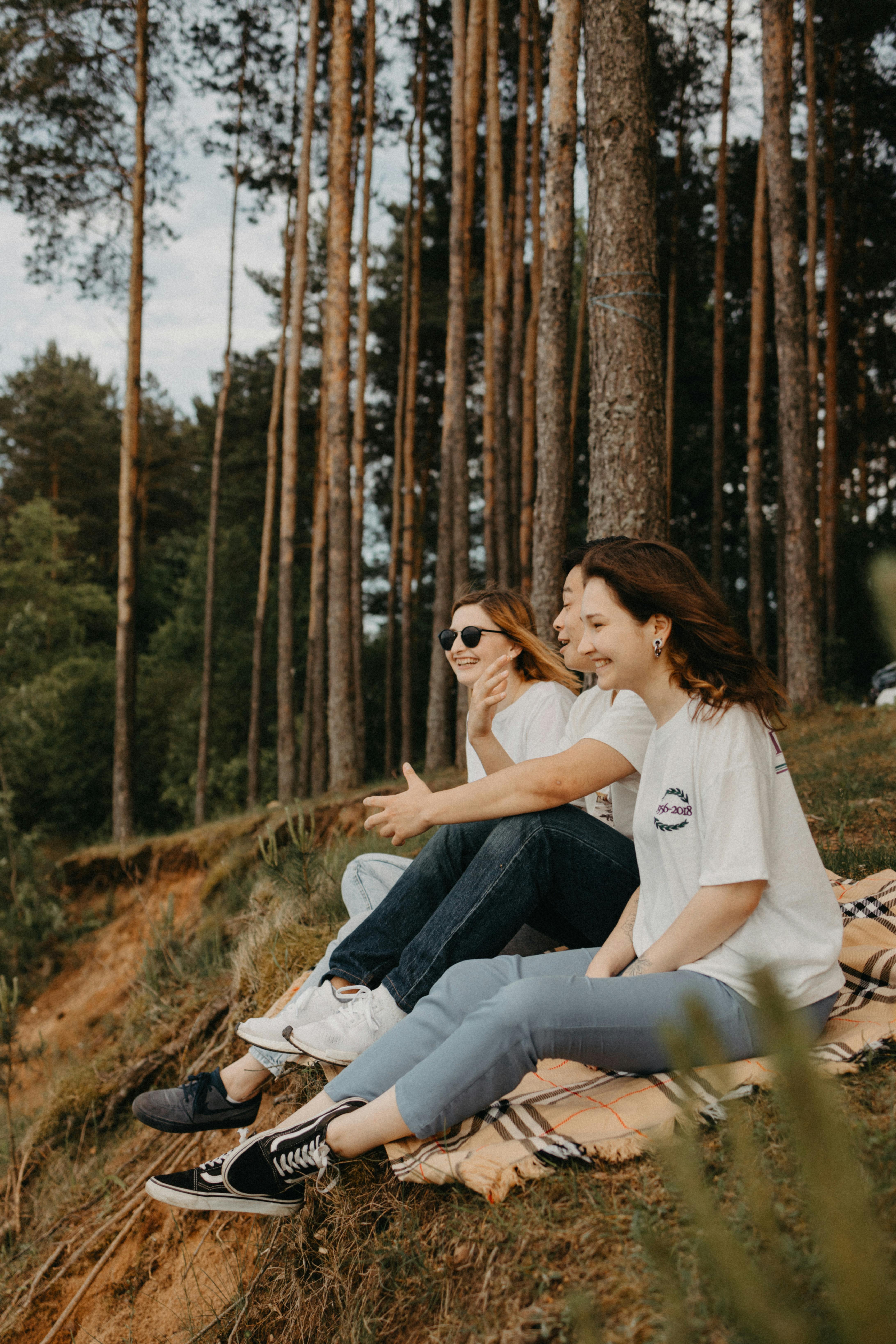 Friends by the Riverbank Two young women sitting on a blanket by a riverbank, surrounded by trees. One is wearing sunglasses and they both are smiling, enjoying the moment.