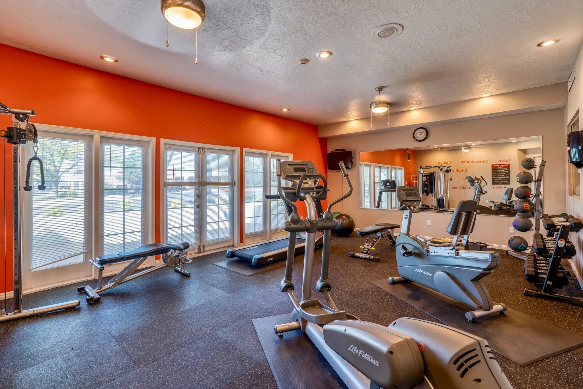 A modern gym interior featuring exercise machines, weights, and large windows with orange walls.
