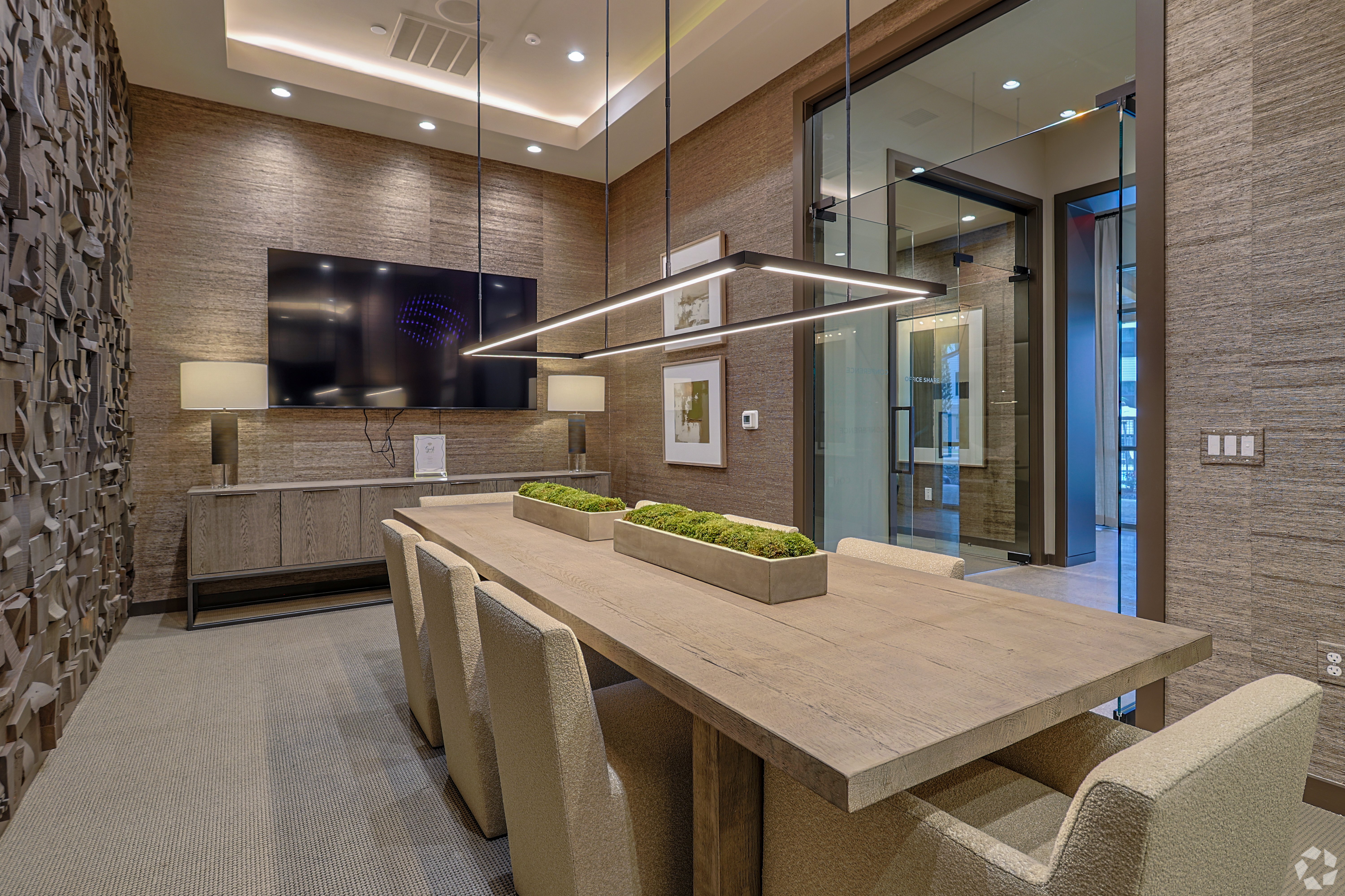 Modern Conference Room Interior A modern conference room featuring a long wooden table with upholstered chairs, a flat-screen TV mounted on the wall, and decorative lighting. Greenery decorates the table, and there are textured wall panels in the background.
