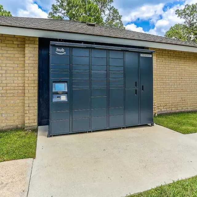 A gray package delivery locker system outside a brick building with green grass around it.