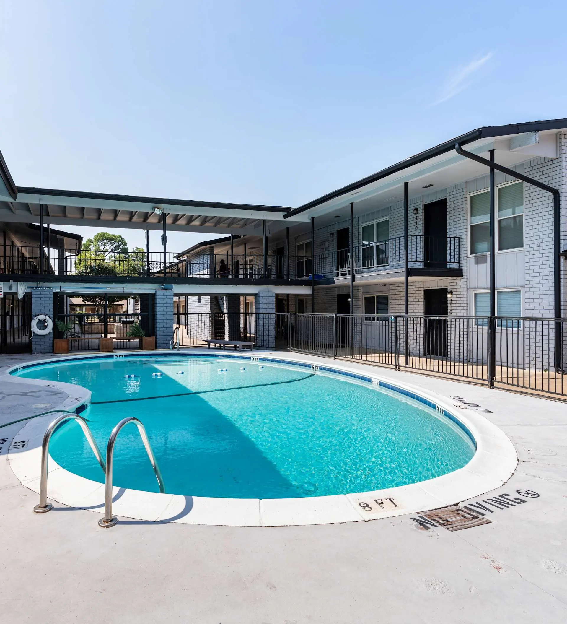 A swimming pool in an apartment complex, featuring clear blue water and surrounded by a fence. Two levels of apartment buildings are visible in the background under a clear sky.