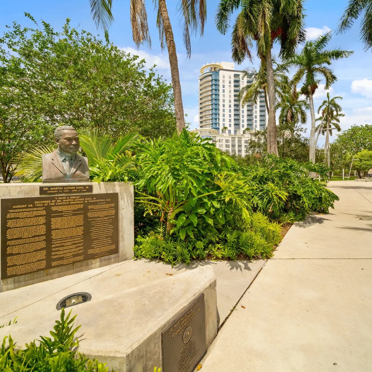 A bust of a man on a stone pedestal, surrounded by lush greenery and palm trees in a public park, with a pathway leading into the distance.
