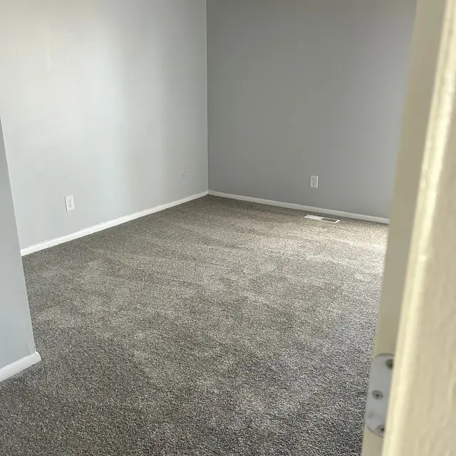 A spacious, empty room with gray carpeting and light gray walls. Wooden beams are visible on the ceiling, and there is a doorframe in the foreground.